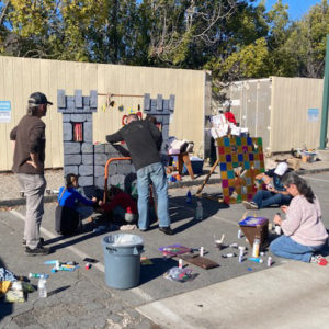 A homemade castle wall and color wall of squares behind a group of adults with tools and painting equipment. There are some tools and paints scattered about the parking lot ground.